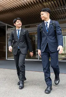 スーツ姿で笑顔を交わしながら歩く二人のビジネスマン、オフィスビルの前でリュックを背負っている